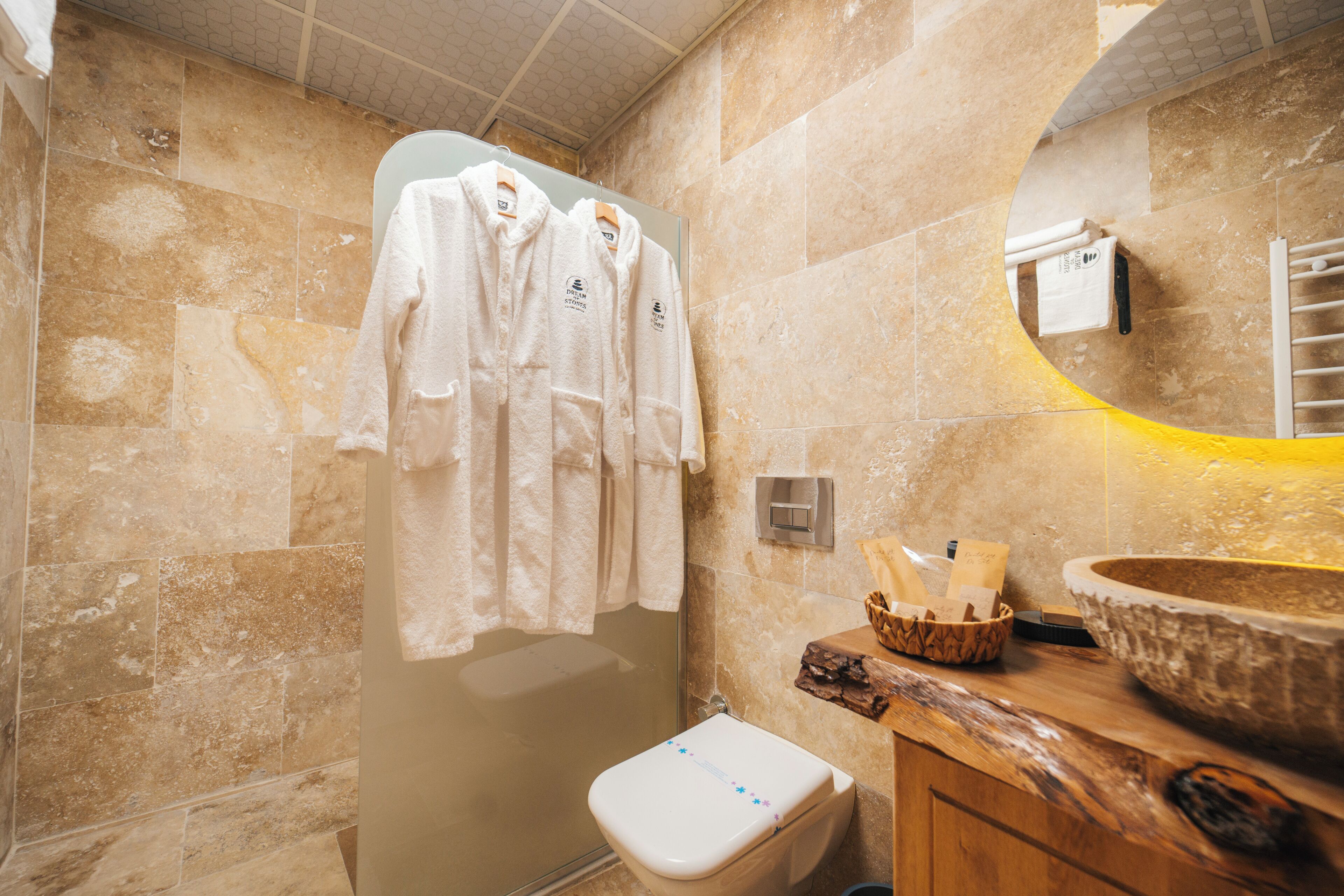 A bathroom with a marble wall, a wooden vanity, a large sink, and three white robes hanging on a towel rack.