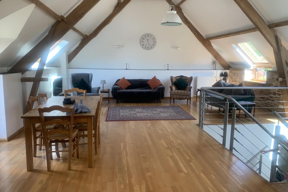 Un loft spacieux avec des poutres apparentes, un coin repas avec une table et des chaises, un canapé noir et un escalier.
