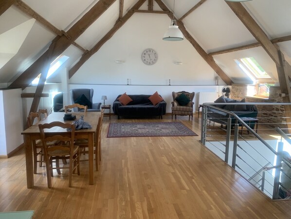 A spacious loft with wooden beams, a dining area with a table and chairs, a black sofa, and a staircase. - Les Rouges Terres Gîtes (Saint-Amand)