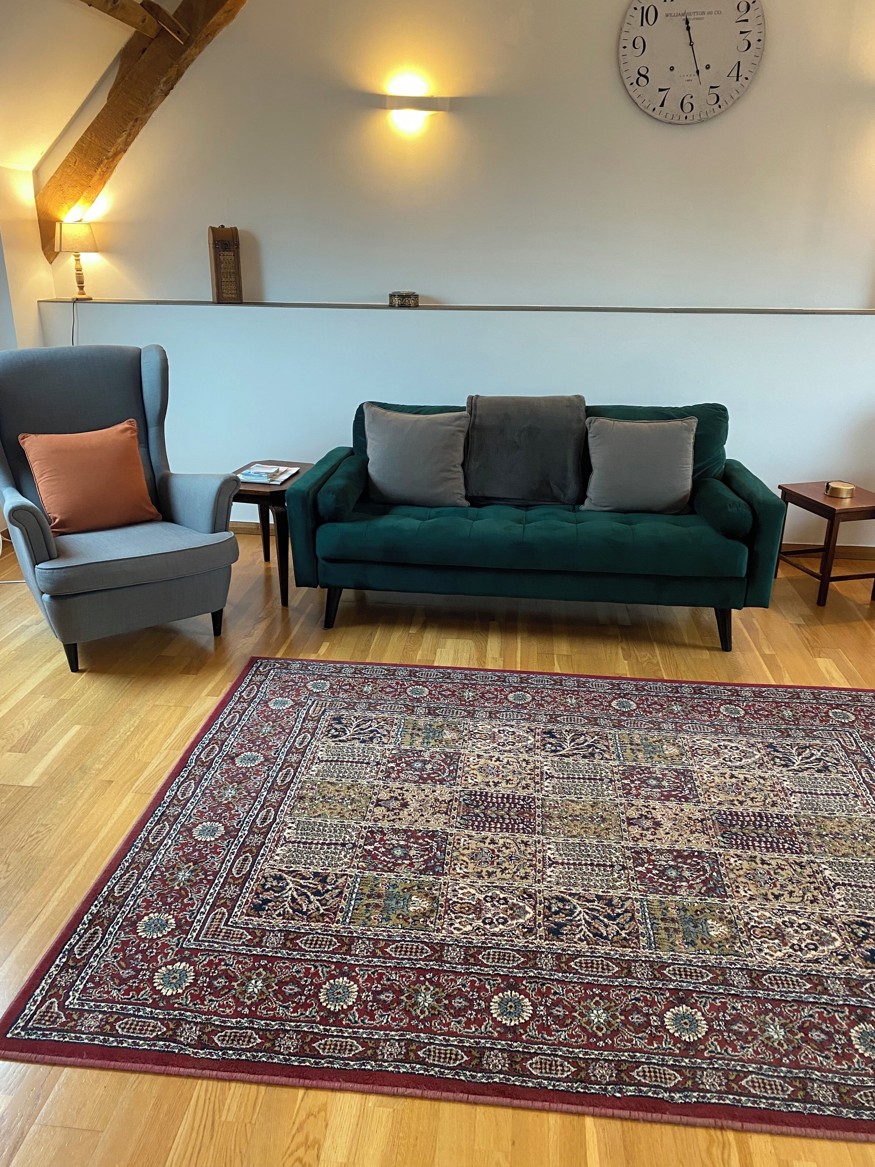 Een woonkamer met een groene bank, een grijze fauteuil, een kamerkleed met dessin, een wandklok en een klein tafeltje.