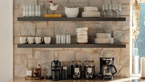 A kitchen with a stone backsplash, shelves with dishes and glasses, a coffee maker, and a cabinet with drawers.