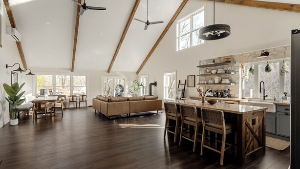 A spacious living area with a wooden ceiling, a large sofa, a dining table, and a kitchen area with a stone backsplash and wooden cabinets.