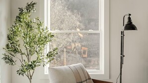 A cozy room with a wooden chair, a small table, a tall floor lamp, and a large window with a view of trees.