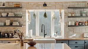 A modern kitchen with a large island, wooden chairs, stone backsplash, and hanging decorations.