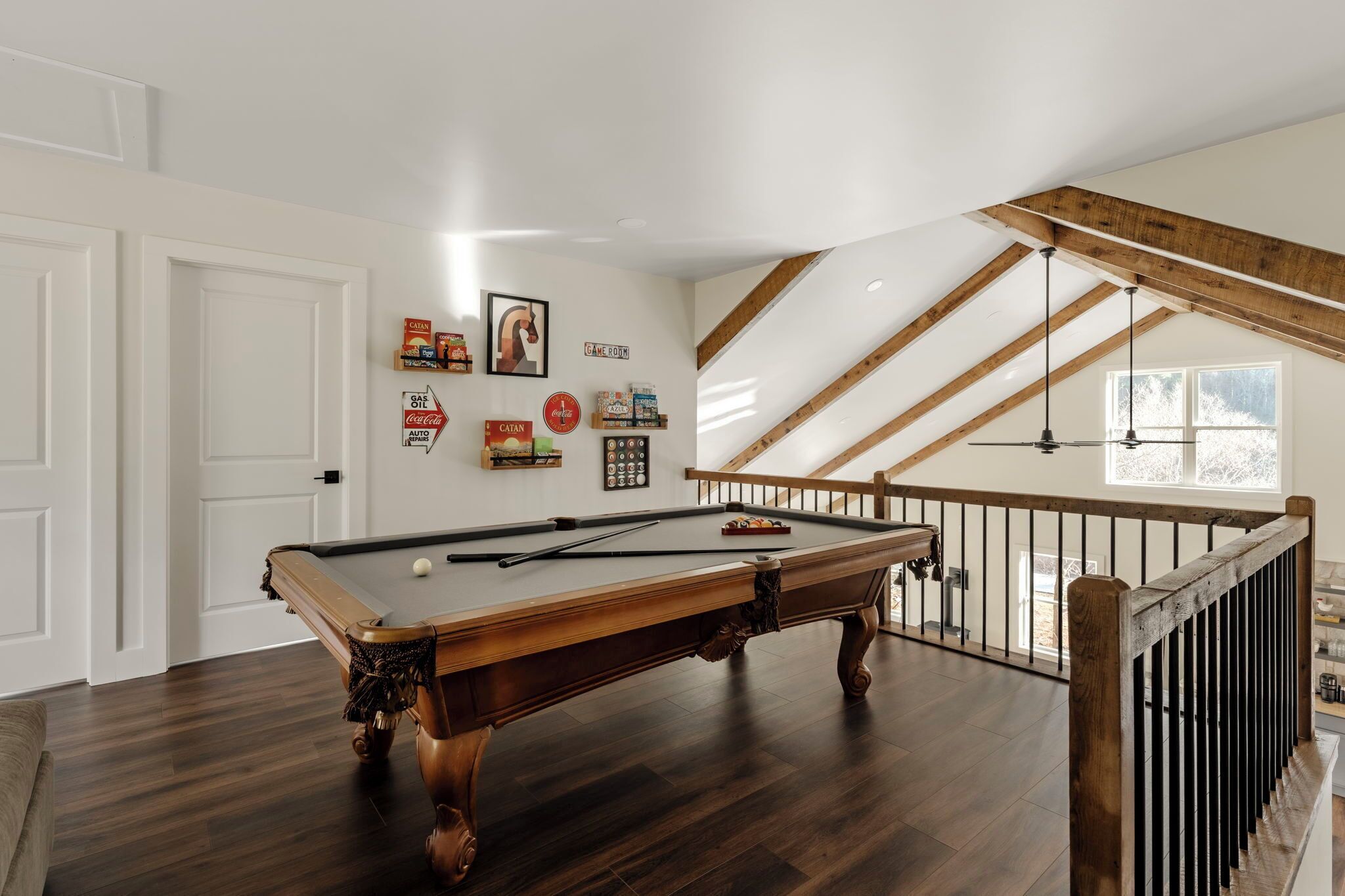 Une table de billard dans une pièce avec des poutres en bois et un plafond en pente, une porte blanche et des éléments décoratifs sur les murs, notamment une horloge et des tableaux encadrés.