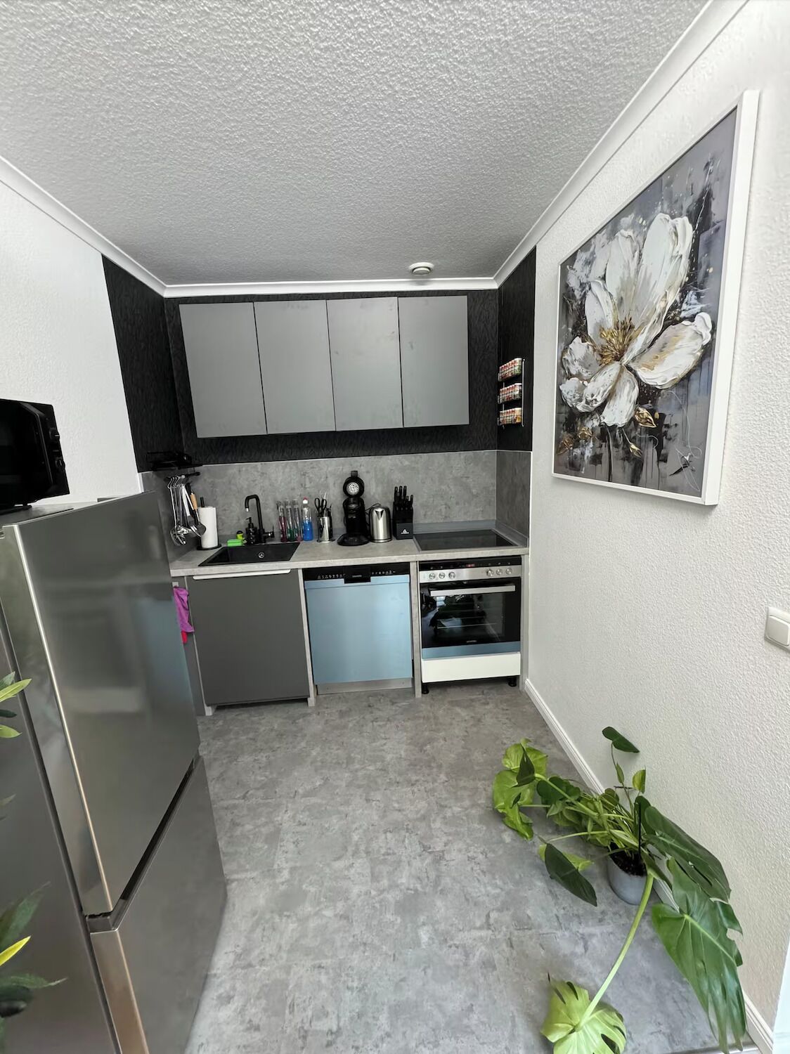 Cocina compacta con gabinetes grises, refrigerador de acero inoxidable, lavavajillas y horno. Hay un fregadero con diversos utensilios de cocina, y un cuadro grande con motivos florales en la pared.