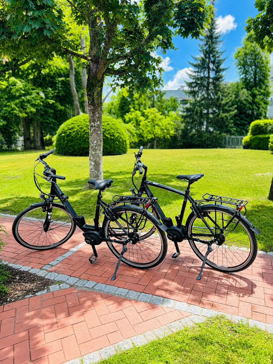 Zwei Fahrräder auf einem Ziegelweg in einem Park mit grünem Rasen und Bäumen.