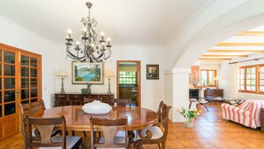 A dining room with a large wooden table, chairs, a chandelier, and a view of the kitchen and living area. - Brisa (Son Servera)