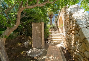 A stone-walled outdoor area with a tiled shower, steps leading to a door, and lush greenery. - Brisa (Son Servera)
