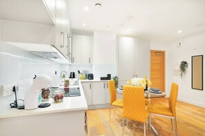 A modern kitchen with white cabinets, a dining area with yellow chairs, and a wooden floor. - Stylish Feltham Studio near Station & Shops 41 (London)