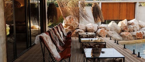 An outdoor patio with a wooden deck, a seating area with chairs and tables, a pool, and a backdrop of rocks and greenery.