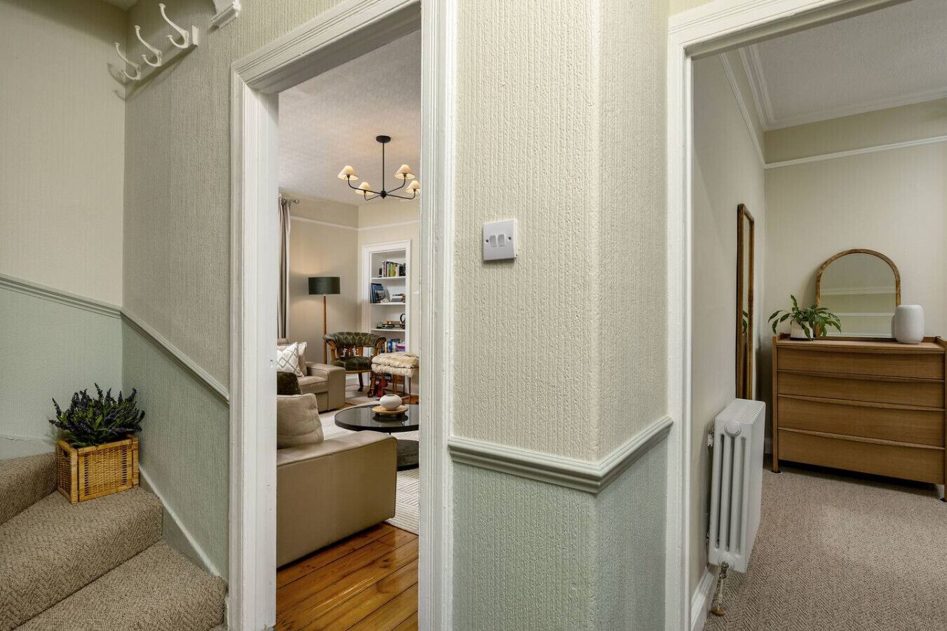 A hallway with a staircase leading to a living room. There is a radiator on the right and a mirror above a wooden dresser.