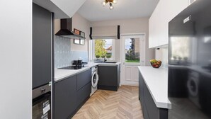 A modern kitchen with a built-in washer and dryer, a herringbone wood floor, and a view of the garden through the window. - Paradykes House - Free Parking & Garden (Midlothian)