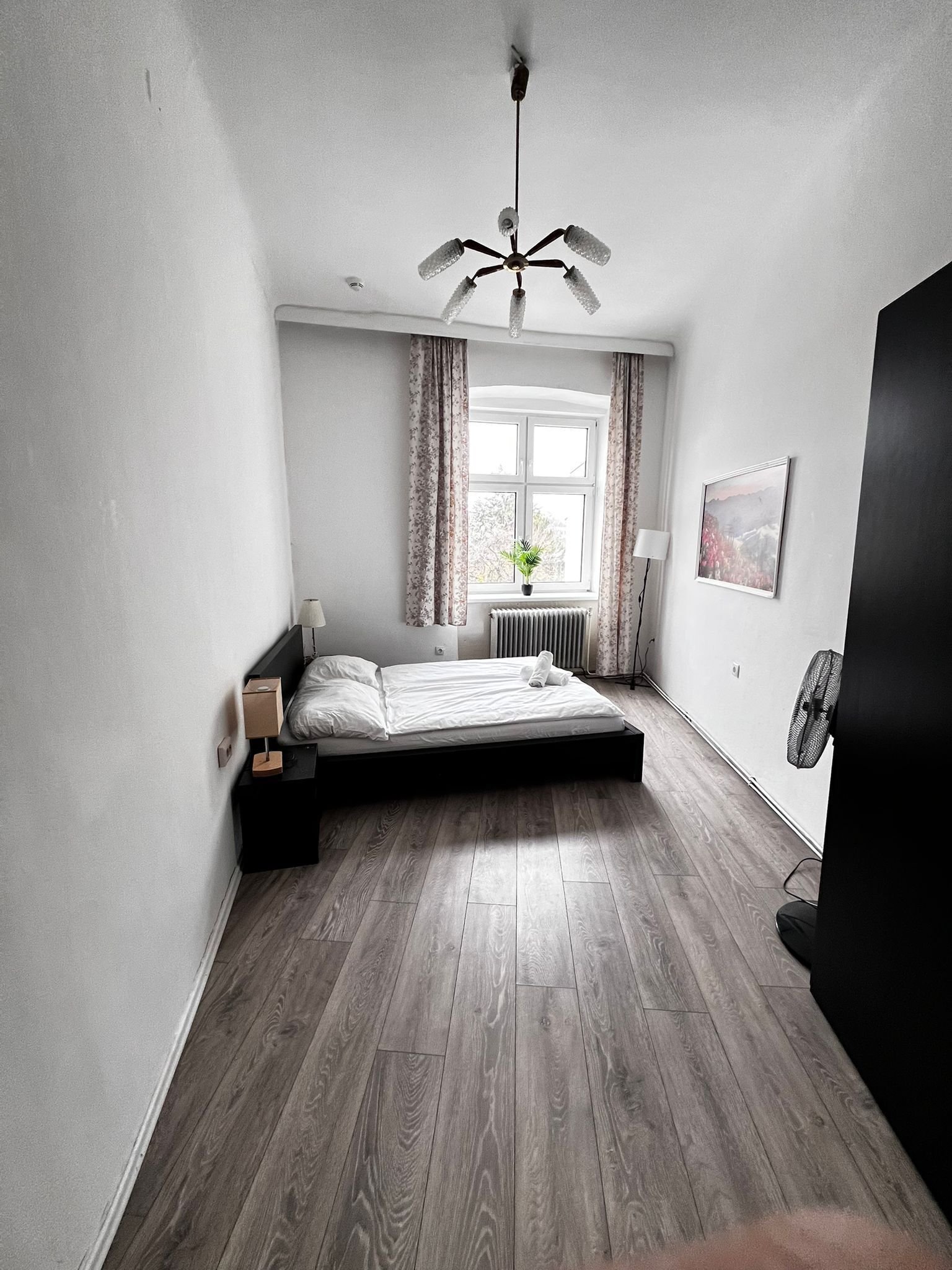 Une chambre à coucher comprenant un lit, un radiateur, une fenêtre avec des rideaux, un ventilateur de plafond et un tableau accroché au mur.