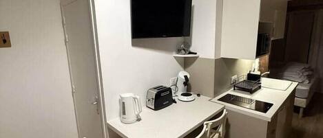 Una cocina compacta con encimera blanca, un lavamanos pequeño, un tostador, una tetera y un televisor de pantalla plana montado en la pared.