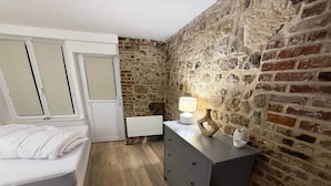 A bedroom with a bed, a dresser, a lamp, and a stone wall. - Apartment in the historic center of Honfleur (Honfleur)