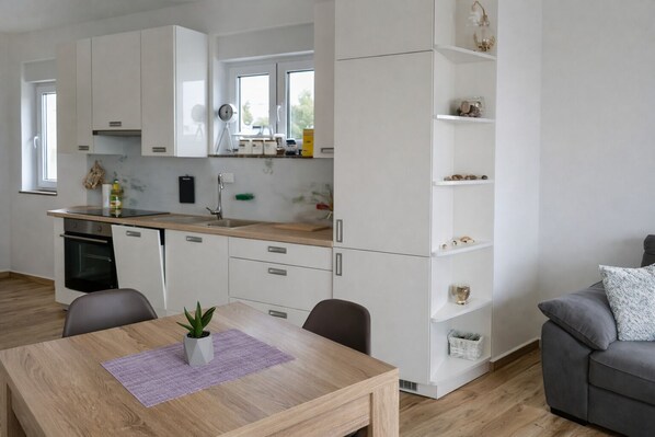 A modern kitchen with white cabinets, a built-in oven, and a dining area with a wooden table and chairs. - Blue Horizon Apartment (Primošten)