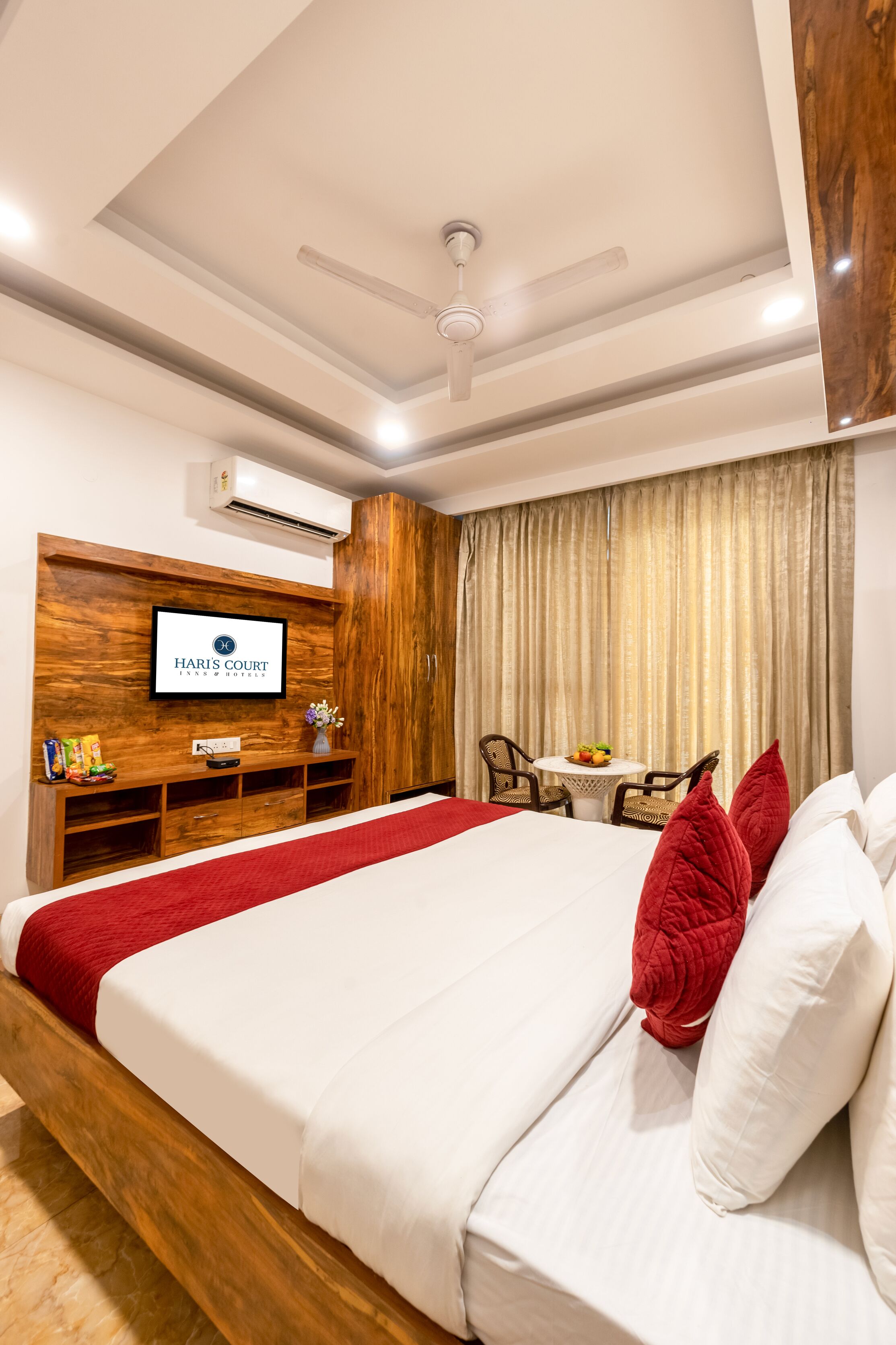 A hotel room with a bed, a television mounted on a wooden wall, a small table with chairs, and a window with curtains.