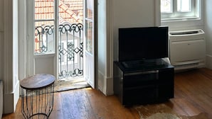 Ein Wohnzimmer mit Fernseher, Holzboden, einer Tür, die auf einen Balkon führt, und einem Fenster mit Jalousien.