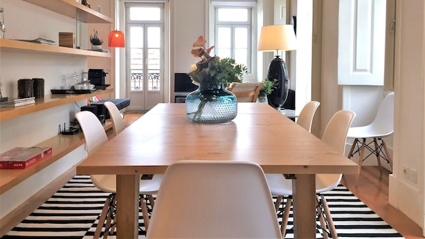 Ein Essbereich mit Holztisch, weißen Stühlen, einem Streifen Teppich und Regalen mit verschiedenen Gegenständen.