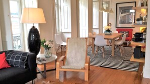 Ein modernes Wohnzimmer mit einer schwarzen Couch, einem weißen Sessel, einem Couchtisch aus Holz und einem Essbereich mit Holztisch und -stühlen.