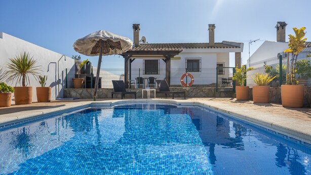 Una piscina con una pérgola, plantas en macetas y un edificio blanco con chimeneas al fondo.