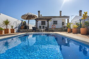 Ein Swimmingpool mit einer Pergola, Blumentöpfen und einem weißen Gebäude mit Schornsteinen im Hintergrund.