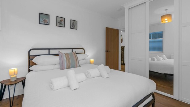Un lit bien fait, avec du linge de lit blanc et des serviettes pliées, une table de chevet avec une lampe, et une porte en bois donnant sur une autre pièce.
