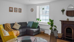Un salon avec un canapé, un fauteuil et une table basse en verre. On y trouve une cheminée et une fenêtre ornée de plantes.