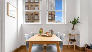 Un coin repas avec une table et des chaises en bois, une plante en pot et une vue sur un bâtiment à travers les fenêtres.