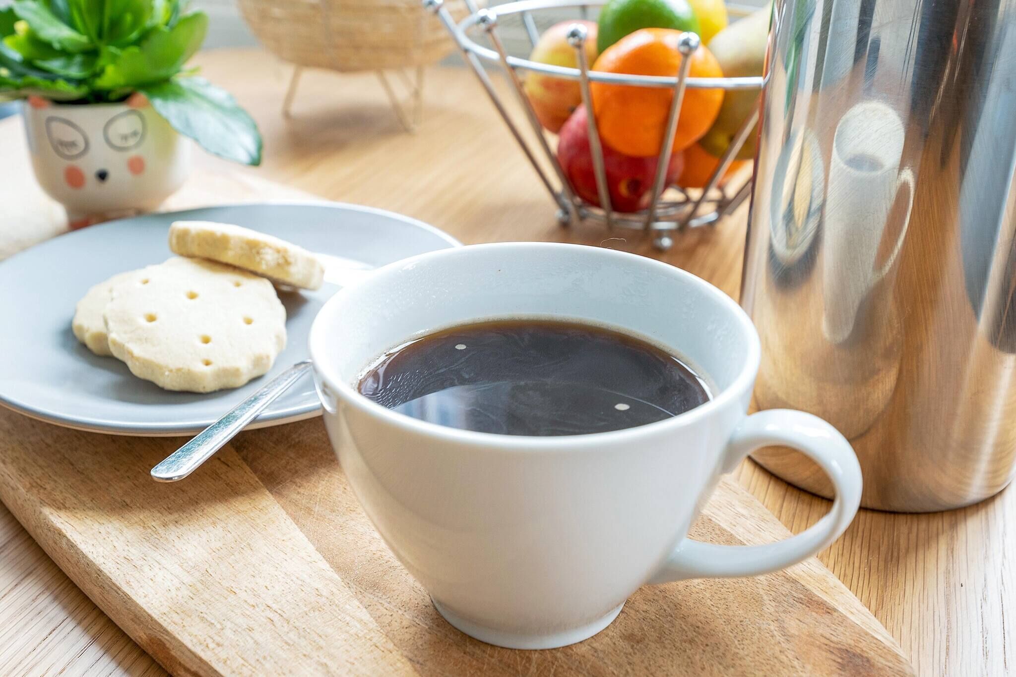 Eine weiße Kaffeetasse auf einem Holztablett, ein Teller mit Keksen, ein Obstkorb aus Metall und ein Metallkrug.