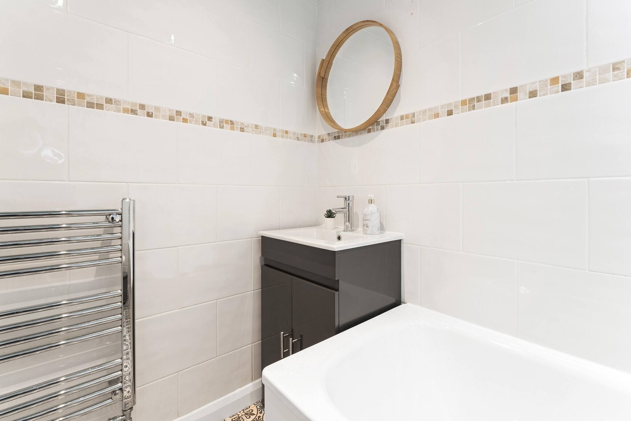 Une salle de bain avec des carreaux blancs, un miroir rond, un lavabo avec des articles de toilette et une baignoire.