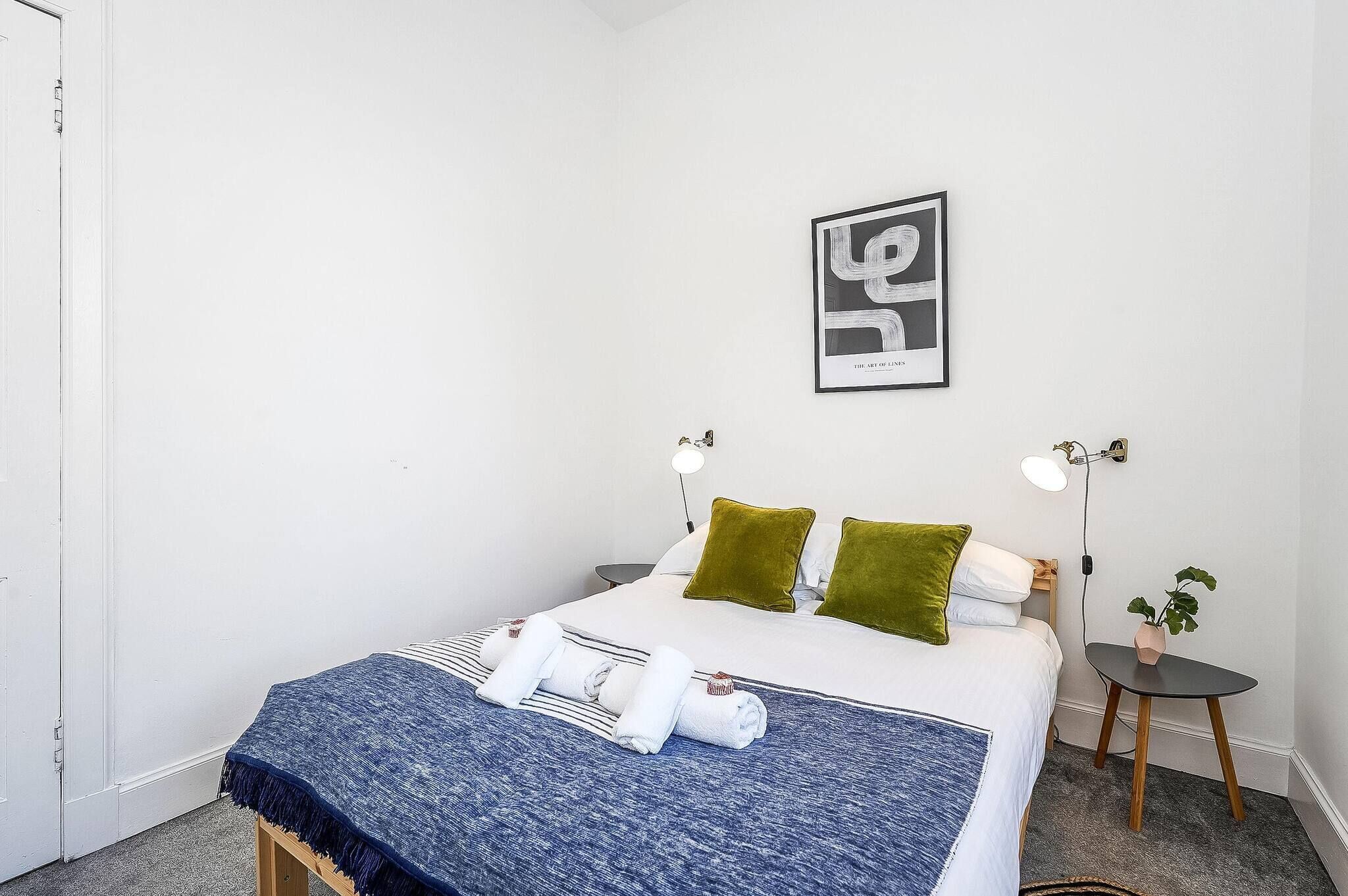 Un lit bien fait, avec du linge de lit blanc et des coussins verts, une couverture bleue et deux tables de chevet avec des lampes.