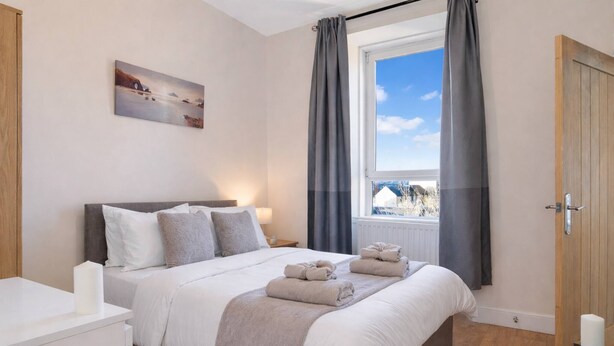 Une chambre à coucher avec un lit, une table de chevet, une fenêtre avec des rideaux, un tableau au mur et une porte.