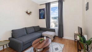 Un salon moderne avec un canapé gris, une petite table basse ronde, une table d’appoint, une plante en pot et une grande fenêtre avec des rideaux.