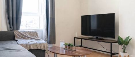 A modern living room with a grey sofa, a wooden coffee table, a flat-screen TV on a black stand, and a potted plant.