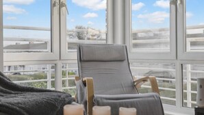Een kamer met een groot raam, een grijze fauteuil en een zwart plaid.