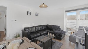Una sala de estar moderna con un sofá de cuero negro, una mesa de centro de vidrio y un comedor con mesa y sillas de madera. Hay cuadros enmarcados en la pared y un ventanal con vistas al exterior.