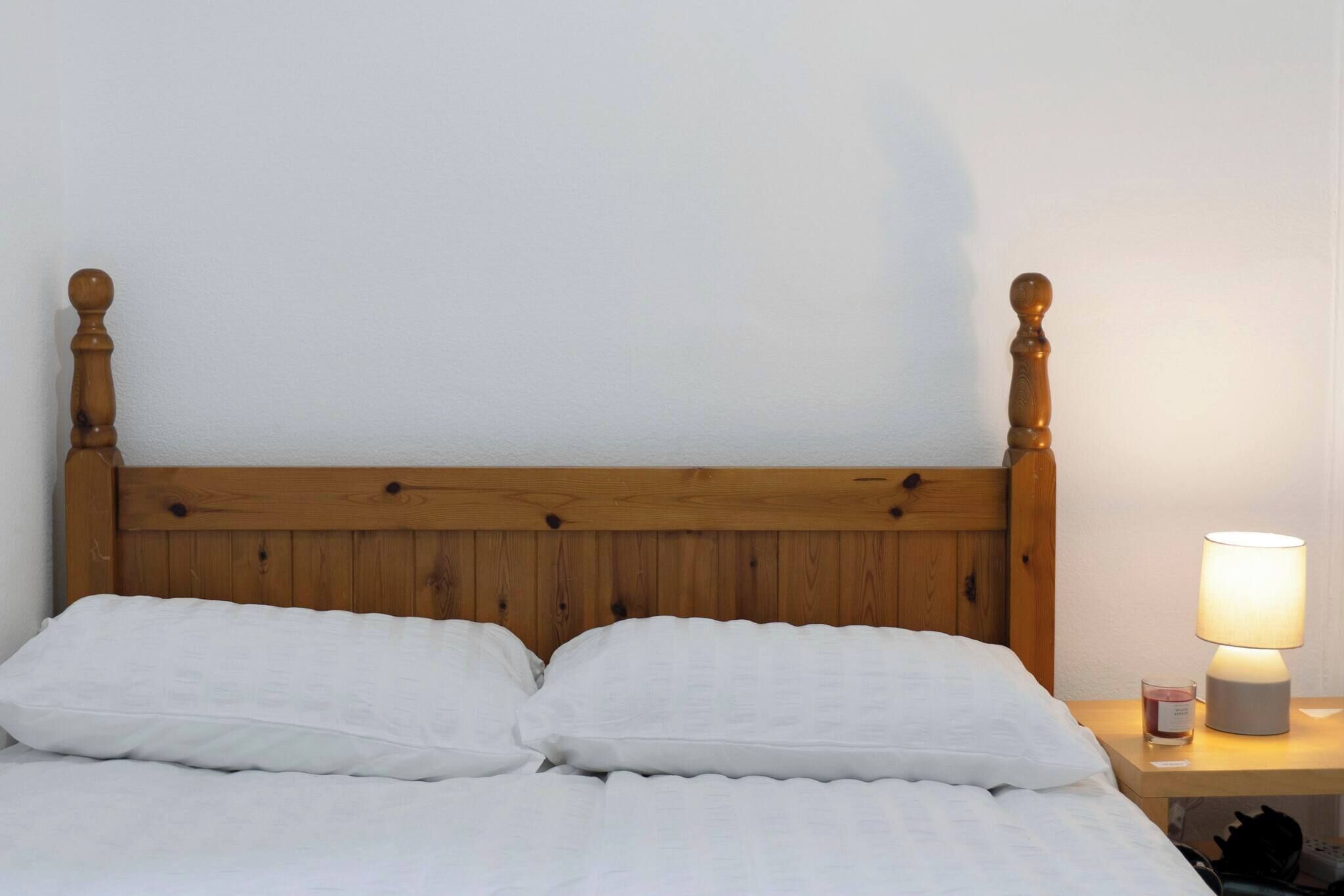 Una cama con cabecera de madera, ropa de cama blanca y una mesita de noche con lámpara y vaso de agua.