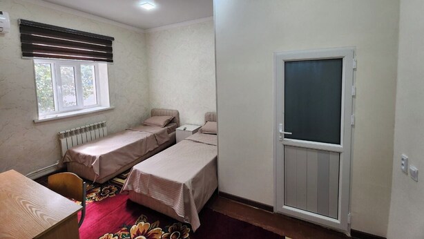 A room with two single beds, a desk, a radiator, and a window with blinds.