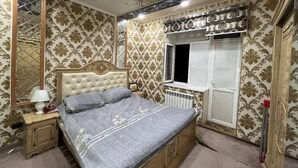 A bedroom with a tufted headboard, a bed with grey bedding, a bedside table with a lamp, a window with a view, and patterned wallpaper.