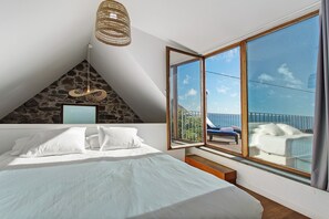 Une chambre à coucher avec un lit, des oreillers blancs, un mur en pierre, une grande fenêtre donnant sur un beau paysage et un luminaire suspendu.