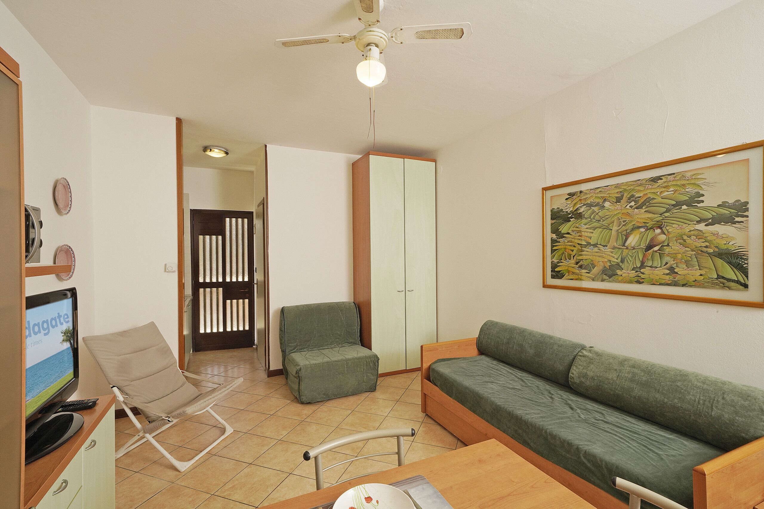 A room with a green sofa, a chair, a small table, a television, and a ceiling fan.