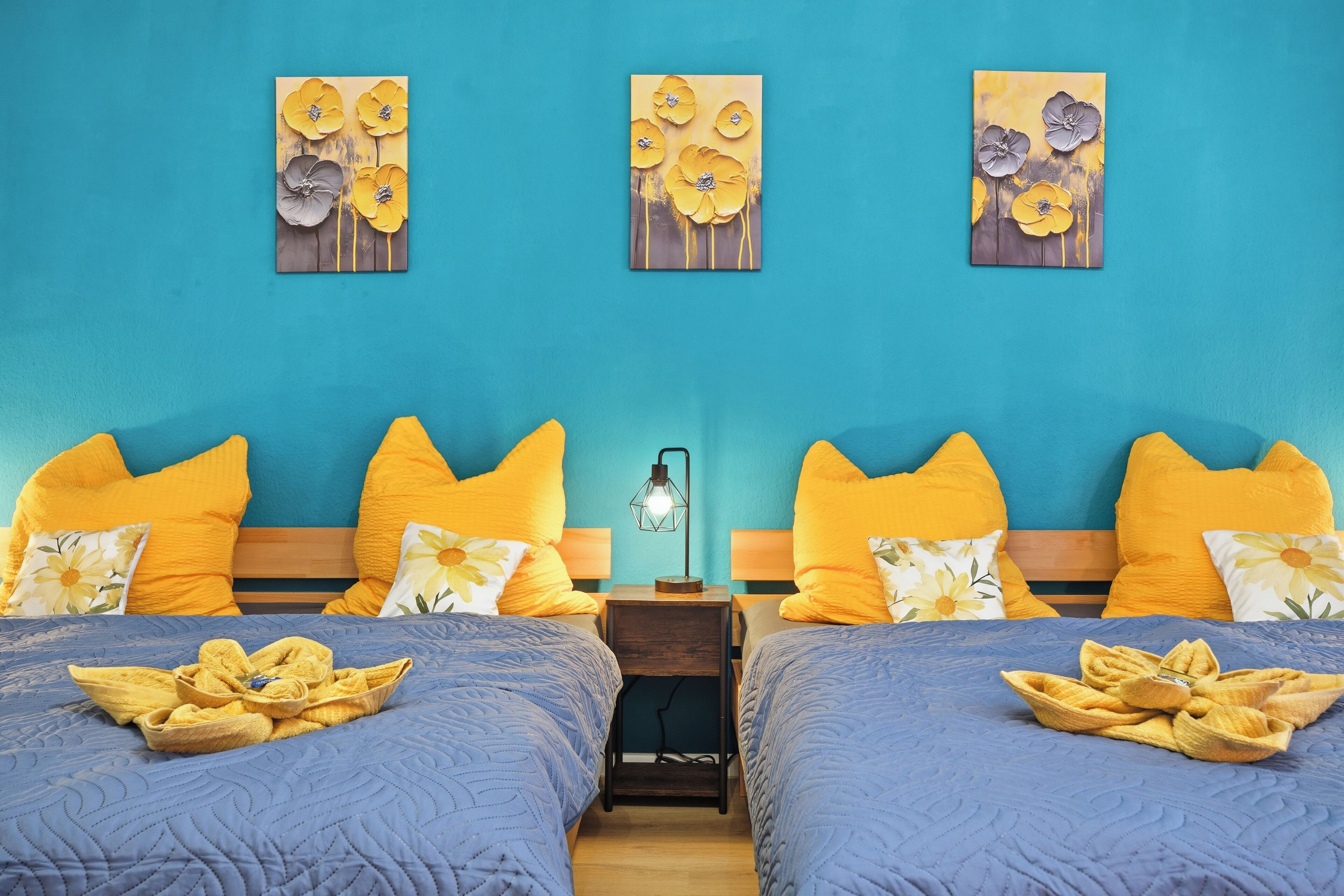 A hotel room with two beds, each with a blue bedspread and yellow pillows. There are three wall-mounted paintings of flowers.