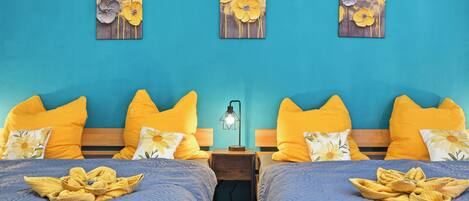 A hotel room with two beds, each with a blue bedspread and yellow pillows. There are three wall-mounted paintings of flowers.
