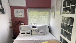 A bedroom with a bed, pillows, a bedside table, a window with blinds, and a wall-mounted air conditioner.