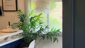 A modern kitchen with a dining area, featuring a coffee-themed wall art, a large potted plant, and a view through the window.