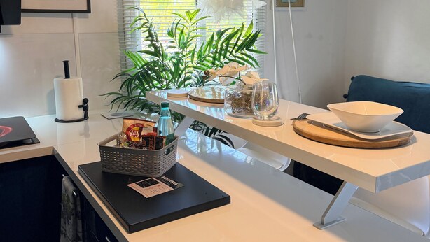 A modern kitchen with a white countertop, a coffee-themed wall art, a potted plant, and a dining area with a bowl and cutlery.