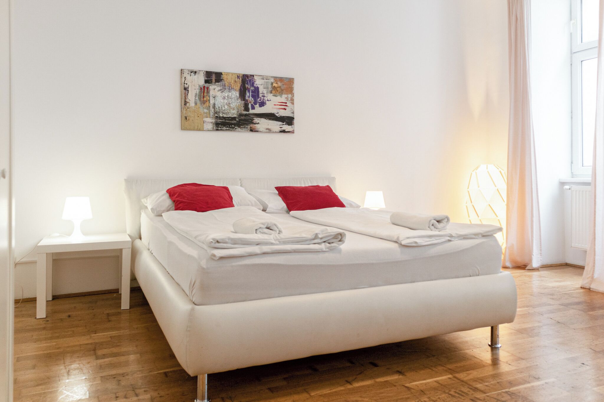 A modern bedroom with a large bed, white bedding, red pillows, and a small bedside table with a lamp.