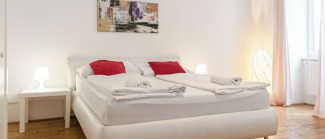 A modern bedroom with a large bed, white bedding, red pillows, and a small bedside table with a lamp.